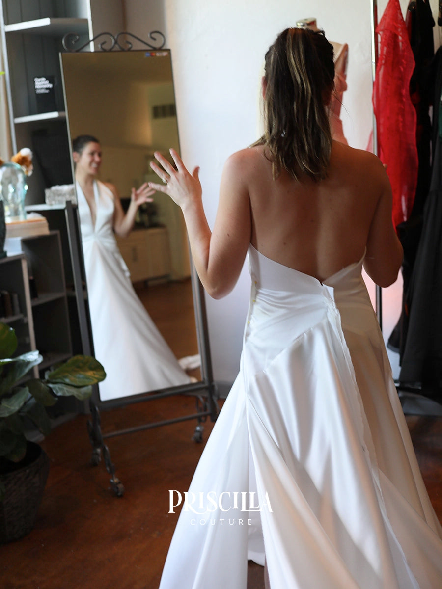 A bride at a fitting session wearing a Priscilla Couture custom white strapless wedding gown with a low open back is seen from behind, standing in front of a mirror that reflects her profile. The dress has a structured satin-like skirt and a draped waist.