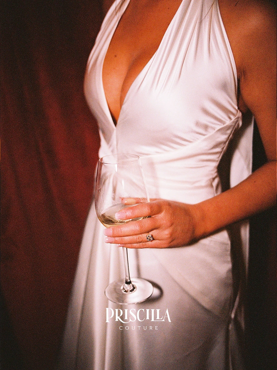 A close-up of a bride wearing a Priscilla Couture custom white silk halter dress and holding a wine glass, with the brand name 'PRISCILLA COUTURE' printed at the bottom.