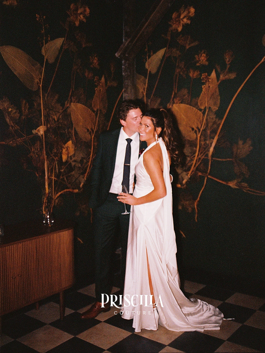 A newly weds embracing in a dimly lit venue with a dark botanical wall mural and checkered floor. The bride wears a Priscilla Couture custom sleeveless white wedding gown with a high slit, and the man wears a black suit with a white shirt and black tie.