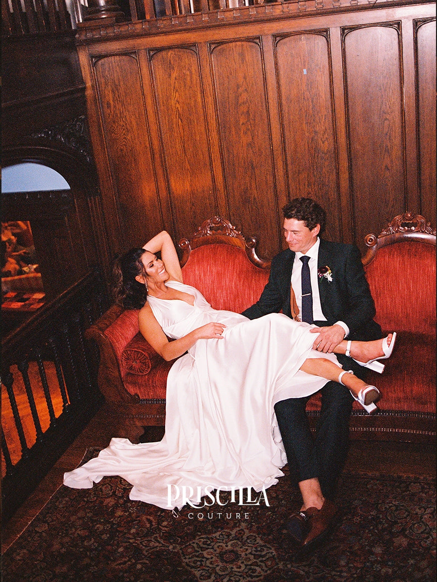 A bride in a Priscilla Couture custom white satin wedding gown reclines on a red velvet sofa while a groom in a black suit holds her up. The photo is taken in a dimly lit room with dark wood paneling, and a 'PRISCILLA COUTURE' watermark is visible at the bottom.