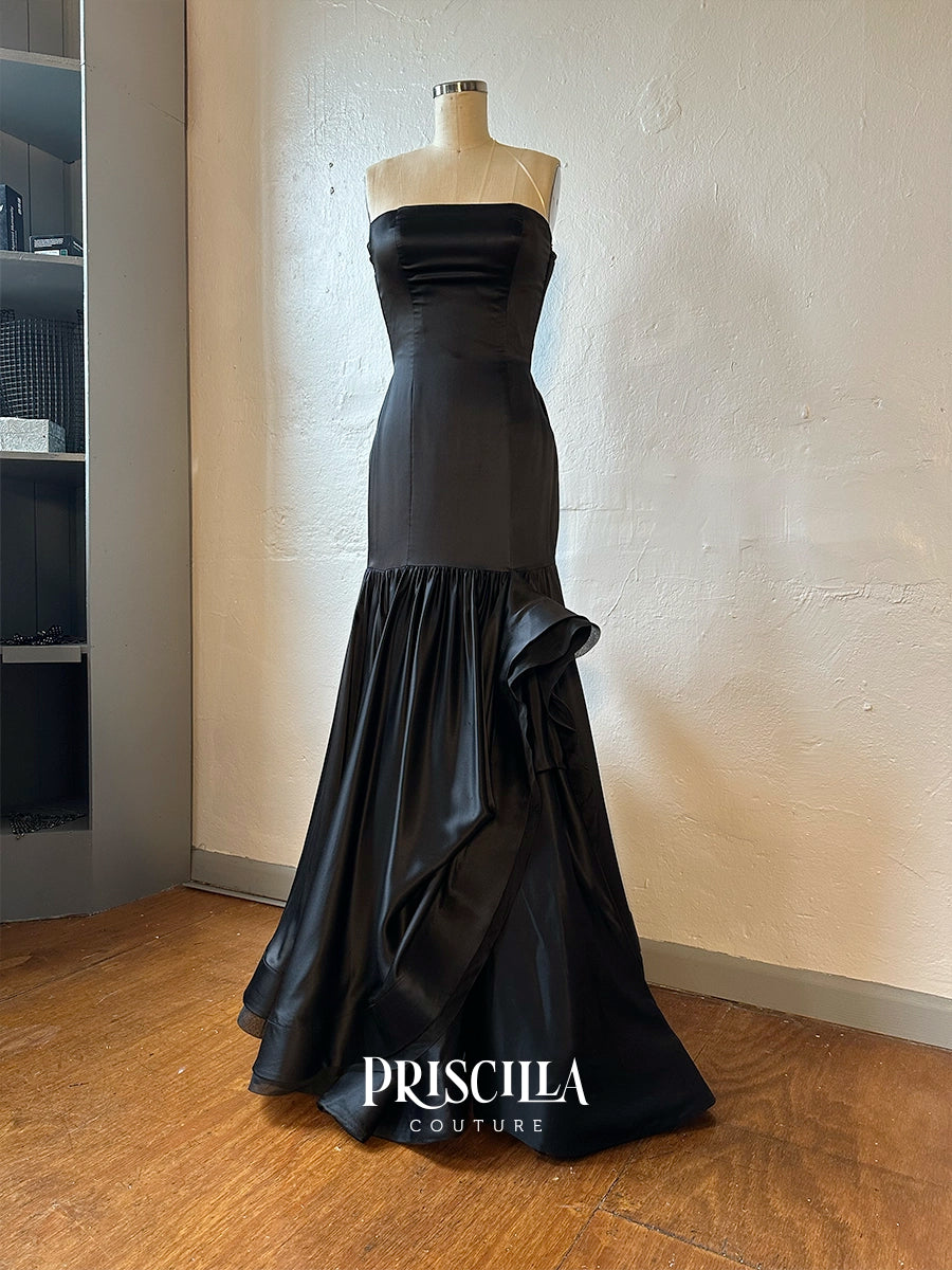 A Priscilla Couture custom strapless black silk evening gown displayed on a mannequin in an indoor room with a textured beige wall and wooden floor. The dress has a fitted bodice and a flared skirt with layered ruffle details.