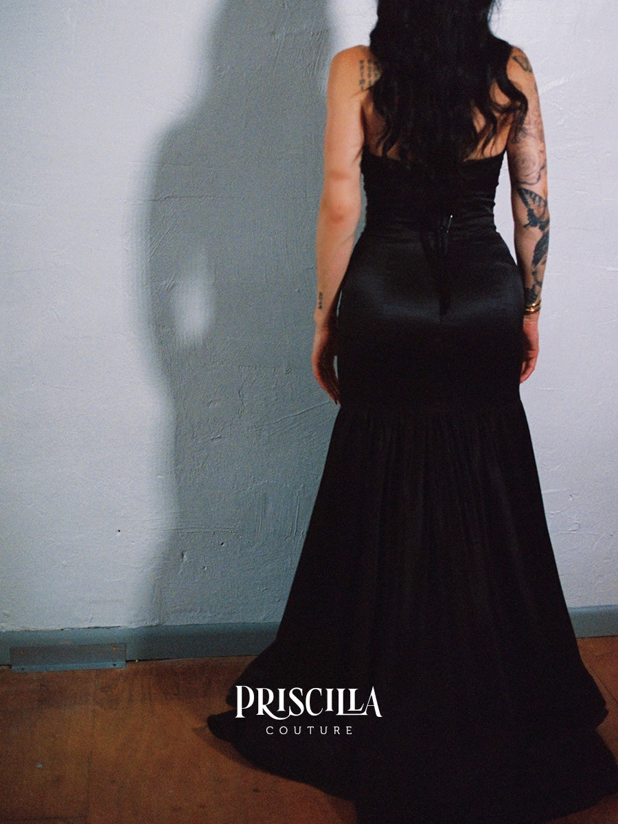 A woman shown from the back wearing a Priscilla Couture custom black silk floor-length gown, standing against a textured light gray wall with a wooden floor. The brand name 'PRISCILLA COUTURE' is printed in white text at the bottom.