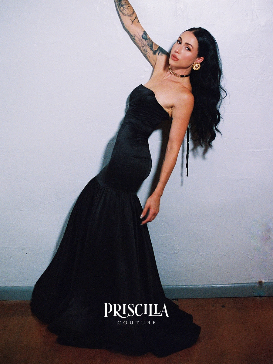 A woman modeling a Priscilla Couture custom strapless black mermaid gown against a textured off-white wall, with the Priscilla Couture logo visible at the bottom.