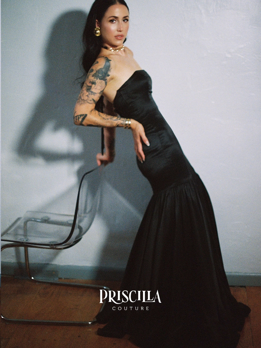 A woman wearing a Priscilla Couture custom strapless black silk mermaid gown poses in a studio setting against a textured light gray wall, with a clear chair to her left and the text 'PRISCILLA COUTURE' at the bottom.