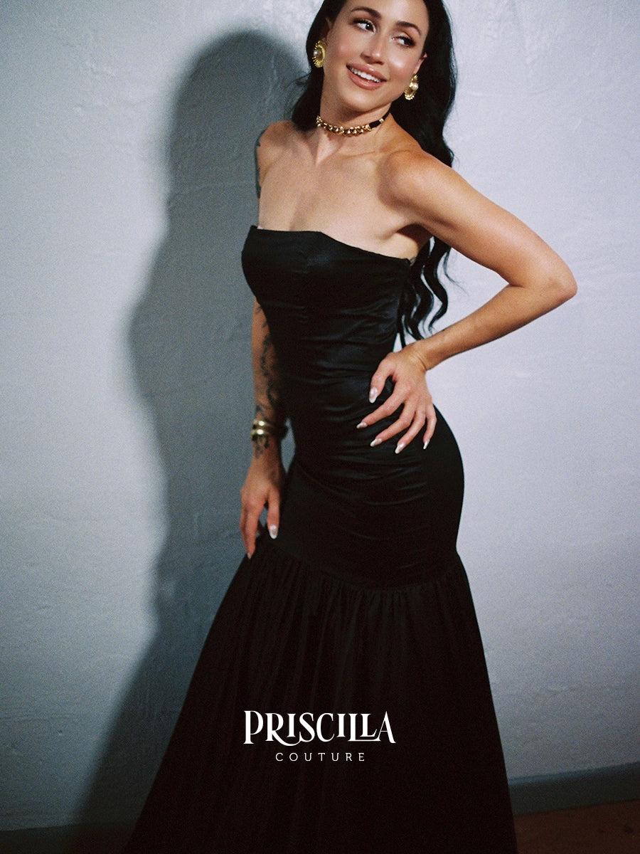 A woman modeling a Priscilla Couture custom strapless black satin mermaid gown against a textured gray wall, with the PRISCILLA COUTURE logo visible at the bottom.