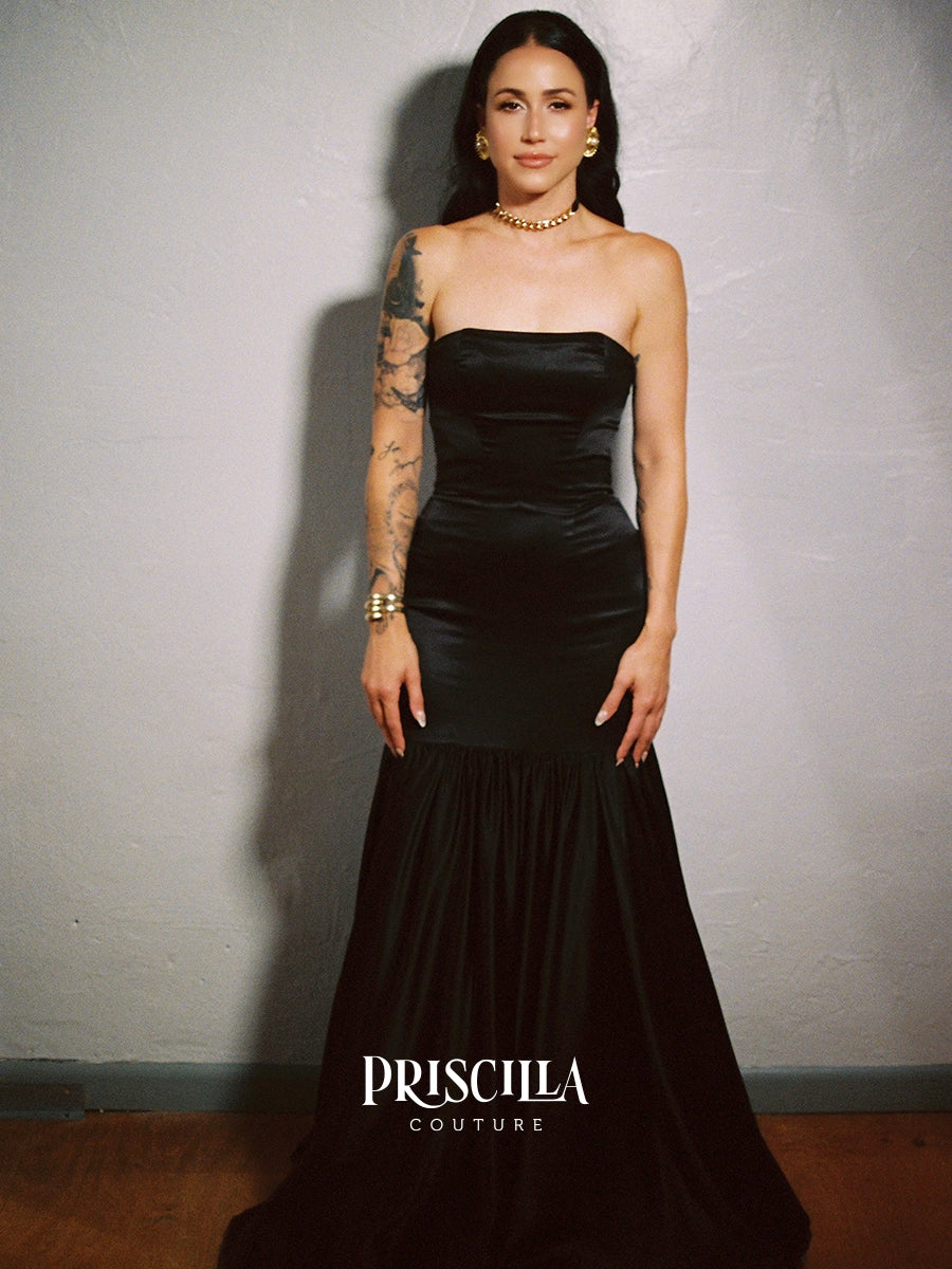 A woman modeling a Priscilla Couture custom strapless black satin gown with a structured bodice and flowing skirt, standing against a textured gray wall. The Priscilla Couture logo is visible at the bottom.