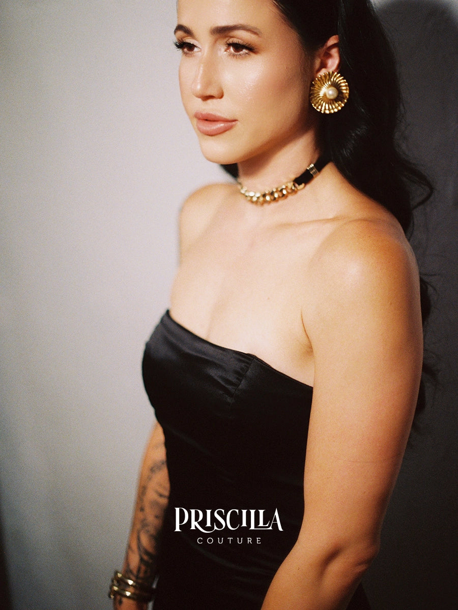 A model in a Priscilla Couture custom black strapless dress wearing gold jewelry, including a chunky gold necklace and a large gold hoop earring, photographed against a dark gray background. The text 'PRISCILLA COUTURE' is visible at the bottom.