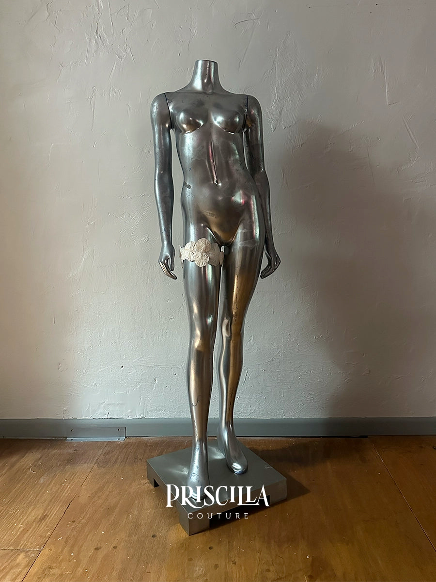 A metallic silver female mannequin torso on a matching base, photographed indoors against a textured light gray wall and wooden floor. The mannequin has no head or arms, with a small white flower garter at the crotch area.