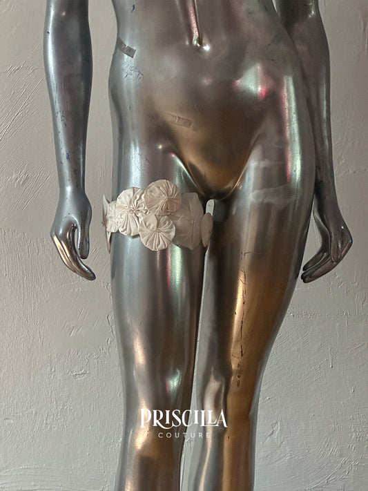 A close-up of a metallic mannequin torso and upper legs wearing a wide cream-colored fabric garter belt with large floral embellishments, set against a textured light gray wall. The brand name 'PRISCILLA COUTURE' is watermarked at the bottom.