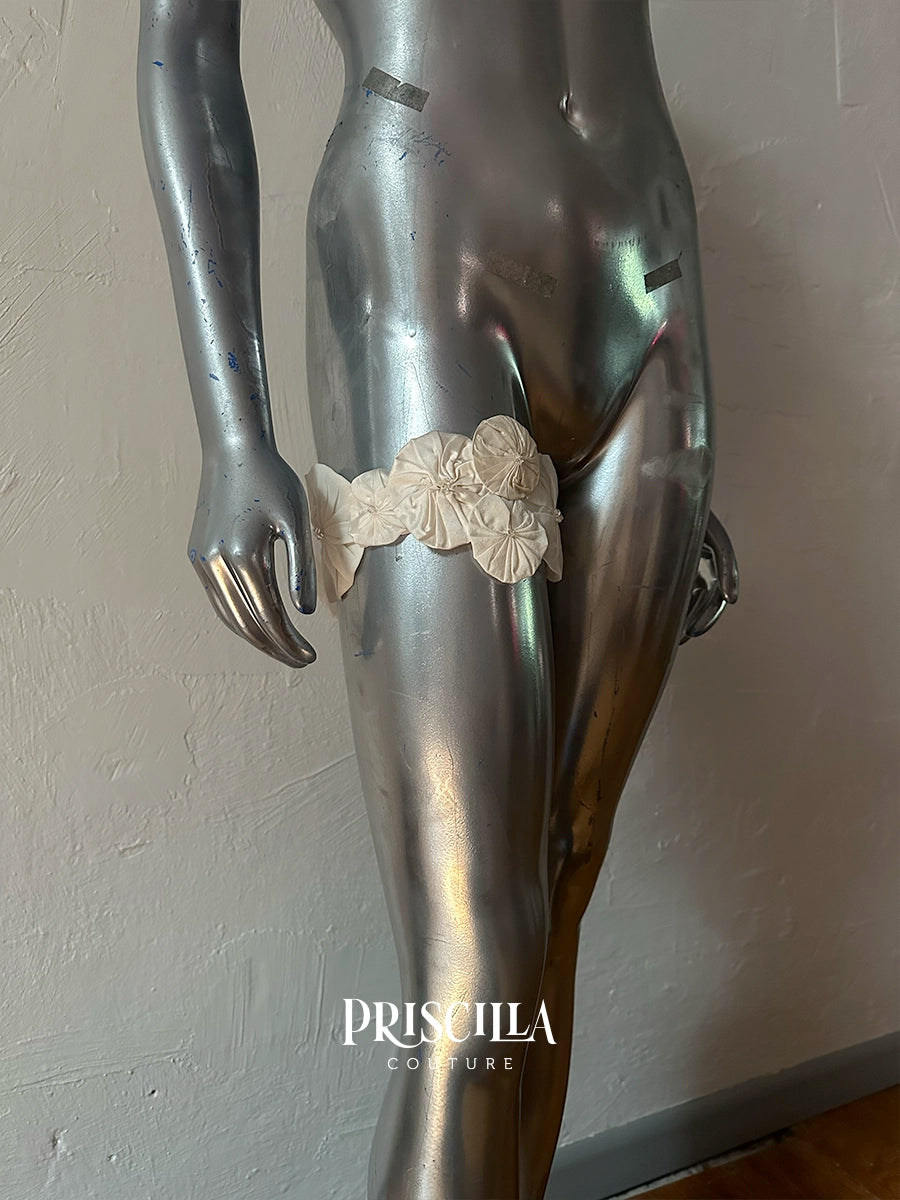 A metallic silver mannequin torso and upper legs wearing a cream-colored fabric Priscilla Couture custom garter belt with floral detailing, set against a textured light gray wall. The brand name 'PRISCILLA COUTURE' is printed in white text at the bottom.