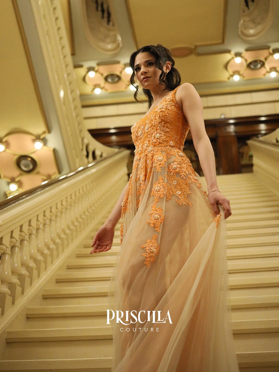 A Priscilla Couture fashion model wearing a Priscilla Couture sleeveless orange gown with intricate floral embroidery and sheer tulle, walking down a grand staircase in a luxurious interior. The brand name 'PRISCILLA COUTURE' is visible as a watermark at the bottom.