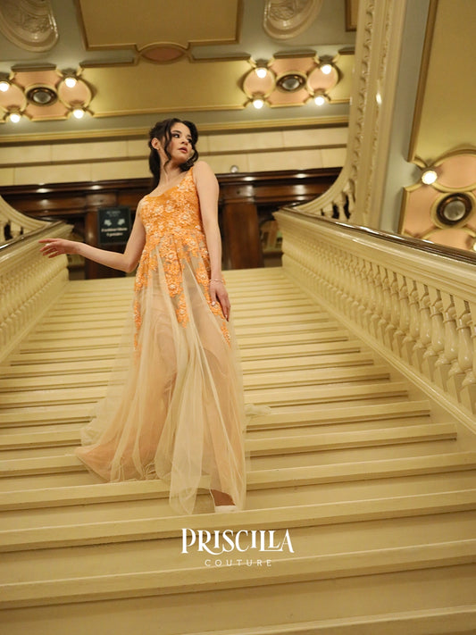 A fashion model wearing a Priscilla Couture sleeveless orange tulle wedding gown with floral applique stands on a grand staircase in an ornate interior. The brand name "PRISCILLA COUTURE" is visible at the bottom.