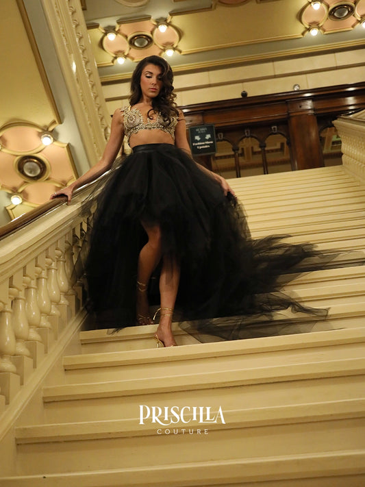 A fashion model wearing a Priscilla Couture custom black tulle two-piece gown with a beaded bralette stands on an elegant indoor staircase. The dress features a voluminous black tulle skirt with a long train, paired with a gold beaded crop top.