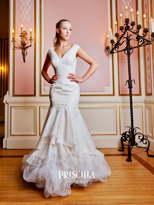 A woman modeling a Priscilla Couture custom white wedding gown in an ornate pink and gold room. The dress has a fitted bodice, a V-neckline, and a tiered mermaid-style skirt.