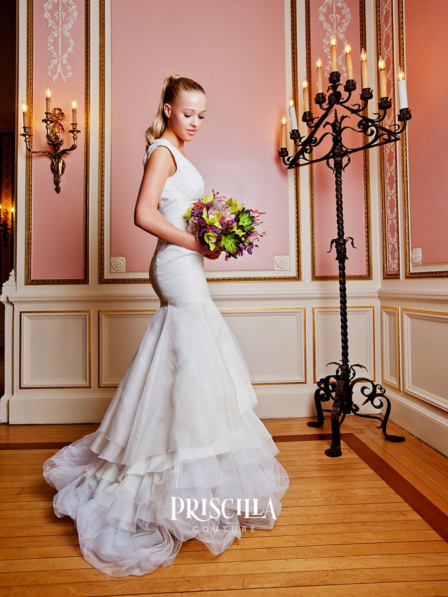 A woman in a Priscilla Couture custom sleeveless white tulle mermaid wedding gown stands in a pink-paneled room holding a bouquet of purple and yellow flowers. A tall black candelabra and wall sconces are visible, with the brand name PRISCILLA COUTURE visible as a watermark at the bottom.