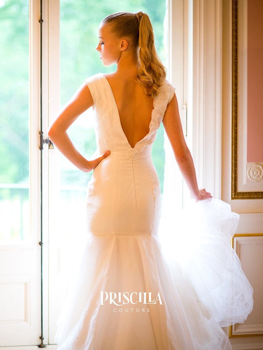 A fashion model wearing a Priscilla Couture custom white wedding dress stands indoors by a bright window, shown from the back. The dress has a deep V back, lace shoulder straps, a fitted bodice, and a full tulle mermaid-style skirt.