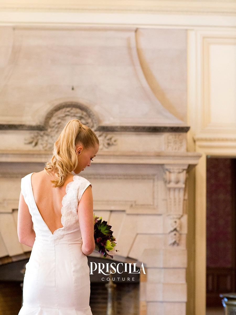 A fashion model in a Priscilla Couture custom white bridal gown with a deep open back and lace shoulder details, standing in front of a large stone fireplace and holding a bouquet. The brand name 'PRSICILLA COUTURE' is watermarked at the bottom.