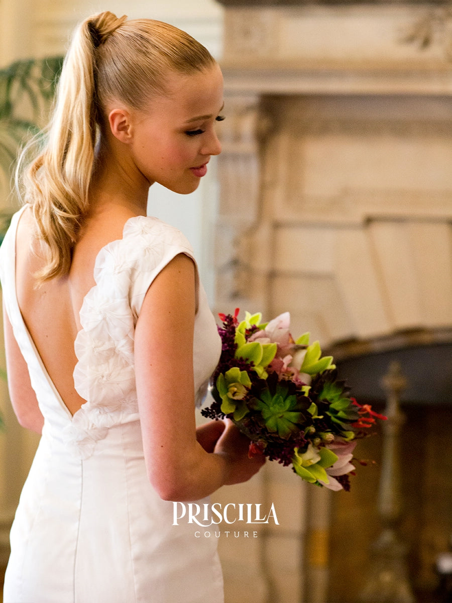 A blonde bride in a Priscilla Couture custom white lace wedding dress with a low open back, holding a colorful bouquet in front of a softly blurred indoor background. The brand name 'PRISCILLA COUTURE' is watermarked at the bottom.