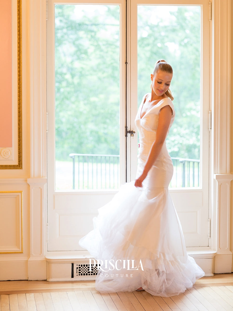 A fashion model wearing a Priscilla Couture custom sleeveless white wedding gown stands indoors in front of tall white French doors, holding out the full train of the dress. The room has warm cream walls and light wood flooring, with greenery visible outside.