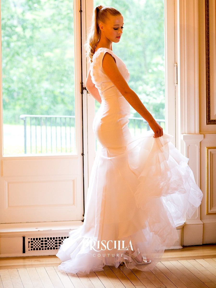 A bride in a Priscilla Couture custom white sleeveless wedding gown stands indoors by large windows, holding out the full train of the dress. The dress has a fitted bodice and a layered, ruffled tulle skirt.