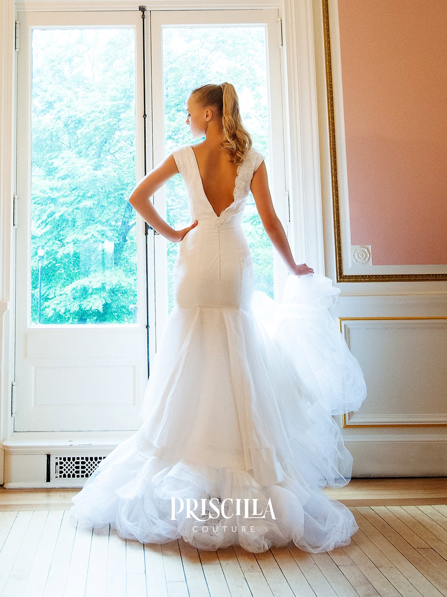 A bride wearing a Priscilla Couture custom white wedding gown stands with her back to the camera in a bright room with tall windows and light wood floors. The dress has a fitted bodice, sheer cap sleeves, a deep V back, and a layered tulle mermaid skirt.