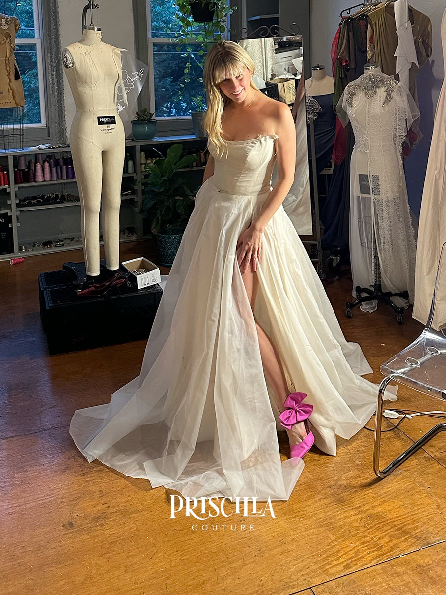 A bride models a Priscilla Couture custom strapless ivory wedding gown with a sweetheart neckline, structured bodice, and a high thigh slit in a bridal boutique. The dress features a long sheer overskirt and train.