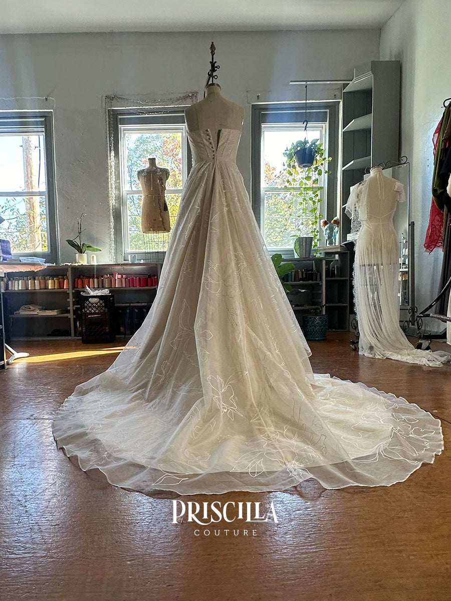 A voluminous Priscilla Couture custom  ivory wedding gown displayed on a dress form in a boutique interior with windows, shelving, and other mannequins. The gown has a fitted bodice, sheer overlay, and a long dramatic train.