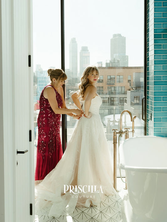 A bride in a Priscilla Couture custom strapless white wedding gown stands by a large window in a modern room, having her back adjusted by a woman in a deep red floral dress. A freestanding bathtub and city skyline are visible in the background.