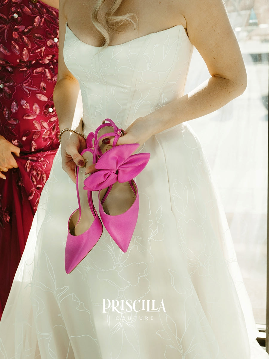 A bride in a Priscilla Couture custom white strapless gown holds a pair of bright pink pointed-toe heels with oversized bows. Another person in a red embellished dress is partially visible at the left edge.