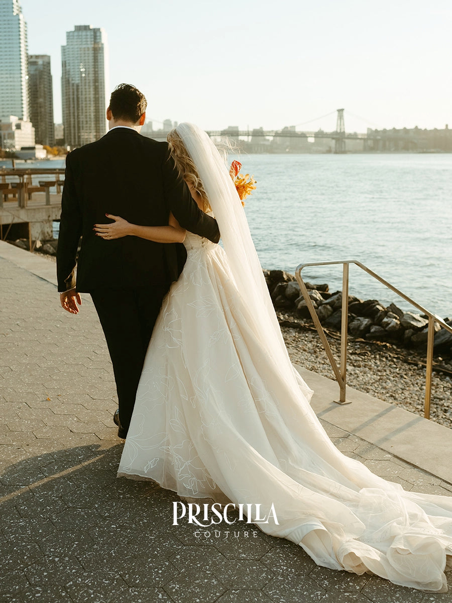 A bride in a Priscilla Couture custom long white wedding gown with a flowing veil and a groom in a black suit walk arm-in-arm along a waterfront promenade, with a city skyline and bridge in the background. The brand name 'PRISCILLA COUTURE' is watermarked at the bottom.