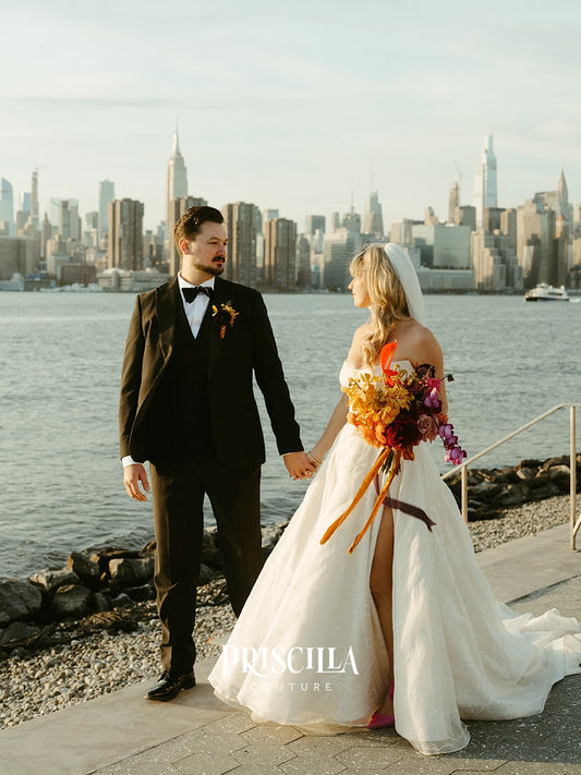 A bride in a Priscilla Couture custom white wedding gown and a groom in a black tuxedo holding hands, standing on a waterfront with a city skyline in the background. The bride holds a large orange and purple floral bouquet.