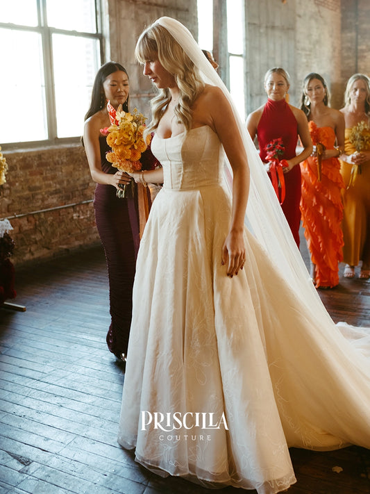 A bride in a Priscilla Couture custom strapless cream A-line wedding gown with a long veil walks indoors while bridesmaids in colorful dresses stand in the background. The Priscilla Couture logo is visible at the bottom.