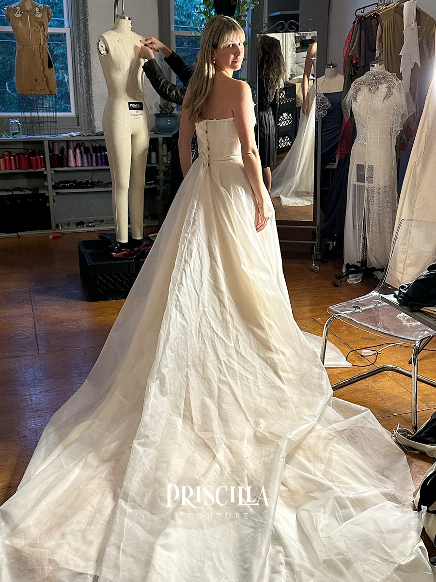 A bride in a fitting session wearing a Priscilla Couture custom strapless wedding gown with a long train, standing in a bridal salon with mannequins and other dresses in the background.