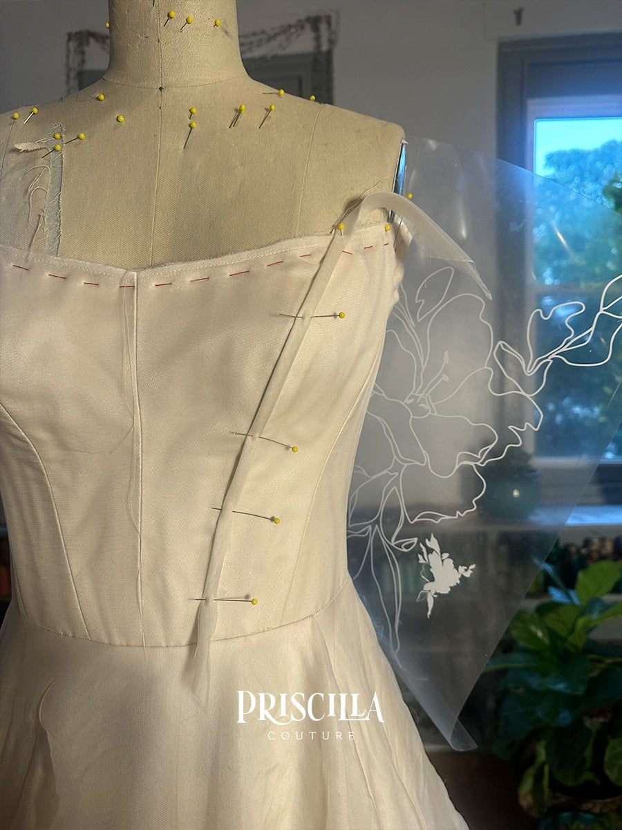 A Priscilla Couture custom cream-colored silk dress on a dress form mannequin, pinned at the shoulders and waist. A sheer white organza overlay with a floral line-art design is draped over the shoulder.