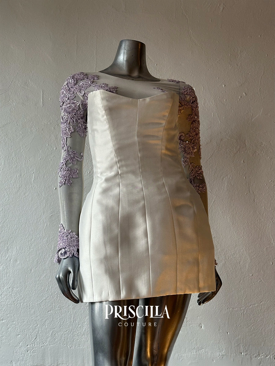 A Priscilla Couture custom strapless silk mini dress in champagne beige with lavender lace long sleeves, displayed on a metallic mannequin against a textured gray wall. The brand name 'PRISCILLA COUTURE' is visible as a watermark at the bottom.