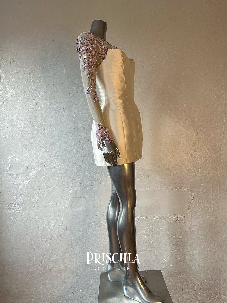A Priscilla Couture custom one-shoulder mini dress displayed on a metallic mannequin. The dress features a cream-colored satin-like body with a lavender-purple floral lace overlay on the long sleeve and shoulder.