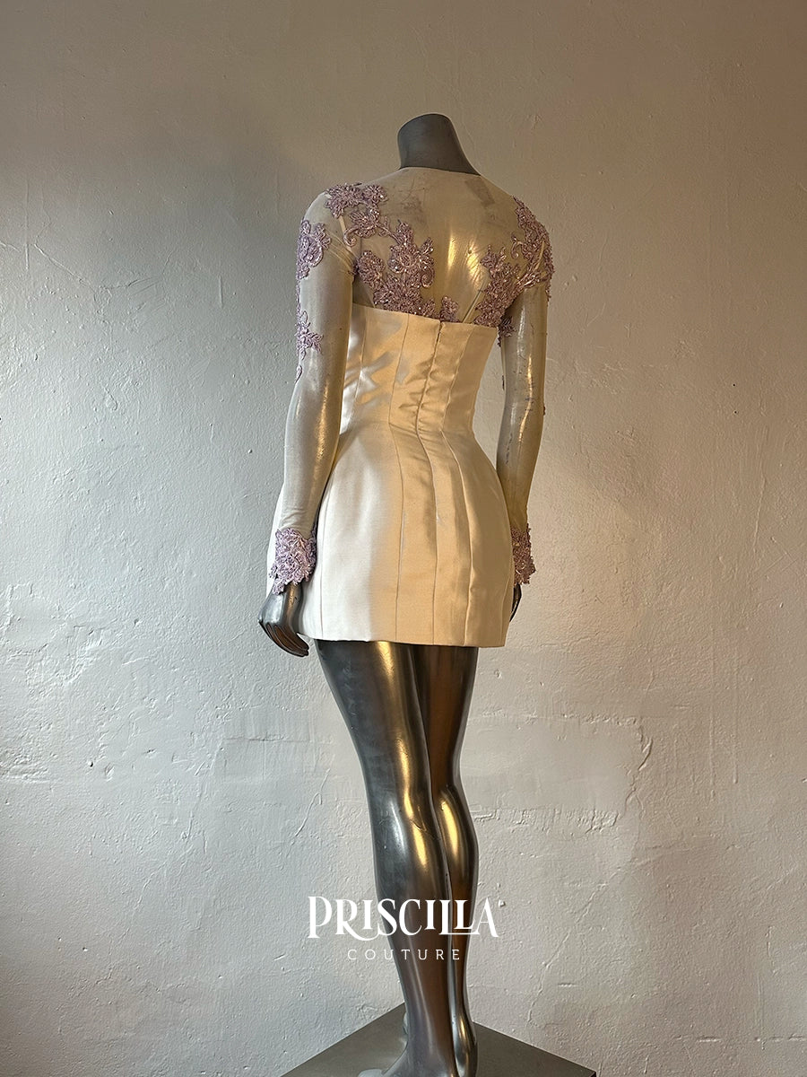 A Priscilla Couture custom champagne-colored mini dress with lavender lace sleeves and a corseted waist, displayed on a metallic mannequin against a textured plaster wall. The brand name 'PRISCILLA COUTURE' is visible as a watermark at the bottom.