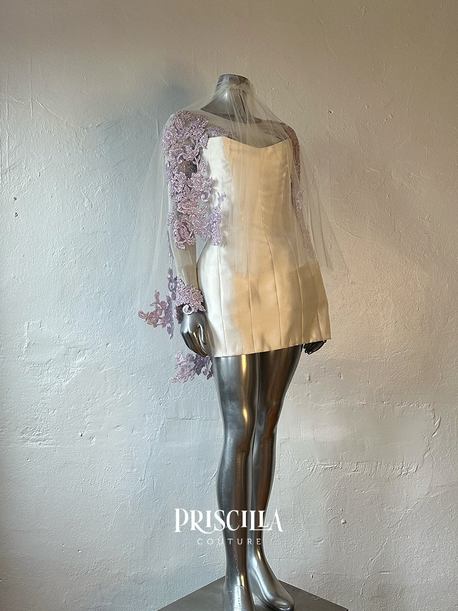A Priscilla Couture custom strapless ivory satin dress displayed on a metallic mannequin, featuring a sheer tulle veil draped over one shoulder with lavender floral lace applique, set against a textured off-white wall. The brand name 'PRISCILLA COUTURE' is visible as a watermark at the bottom.