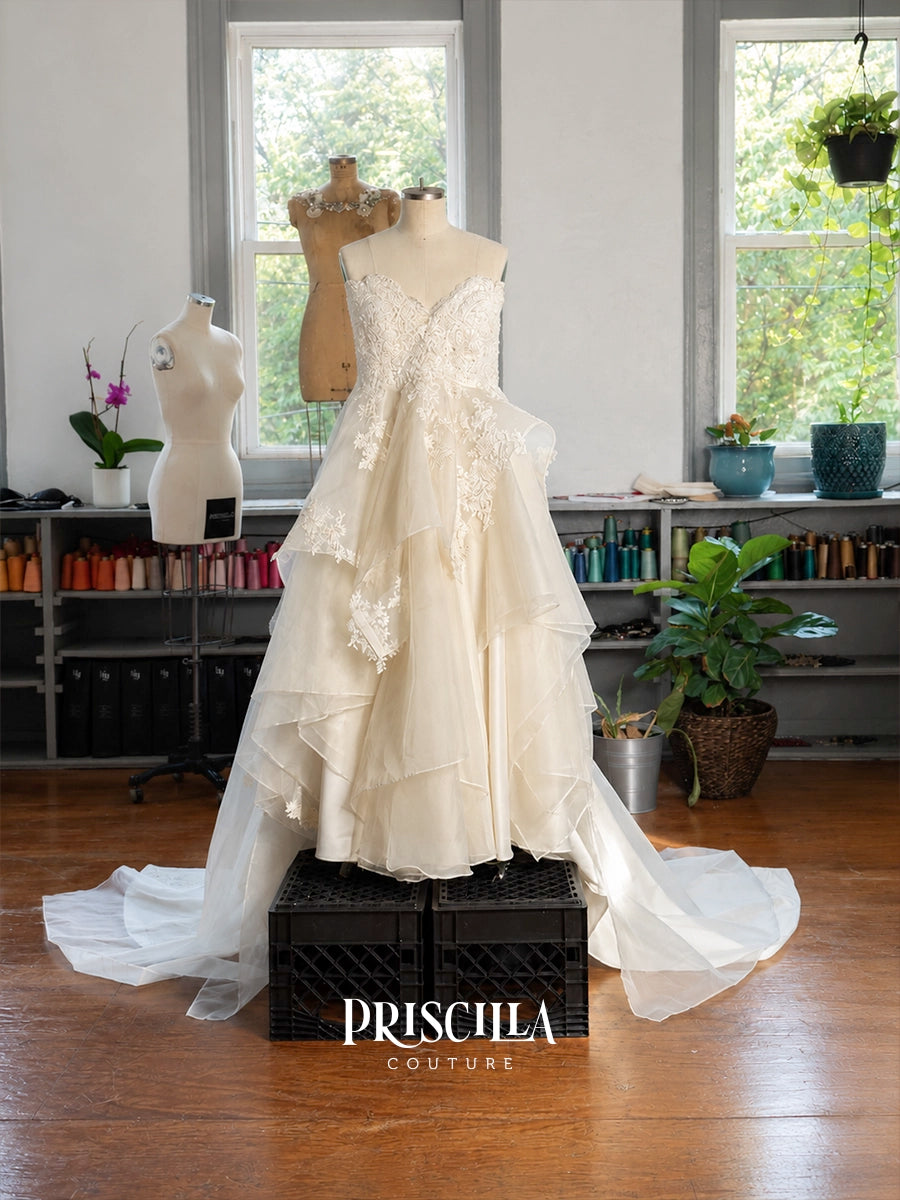 An ivory A-line Priscilla Couture custom wedding gown with lace applique and a layered ruffled skirt is displayed on a mannequin in a bridal studio. The dress sits on a black crate with the brand name 'PRISCILA COUTURE' printed at the bottom.