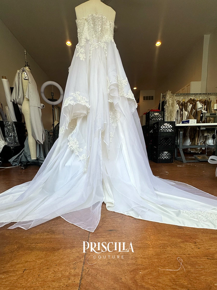 A Priscilla Couture custom white strapless wedding gown displayed on a mannequin in a bridal boutique. The dress features a heavily embellished lace bodice, layered tulle overskirts with floral applique, and a long train.