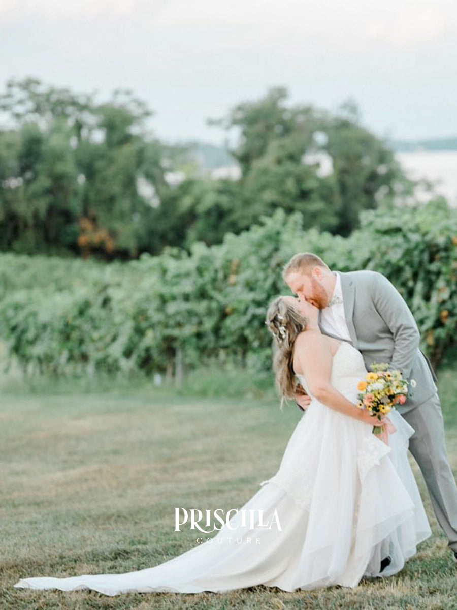A bride in a Priscilla Couture custom white wedding gown and a groom in a light gray suit kiss in a vineyard. The bride holds a yellow and orange floral bouquet.