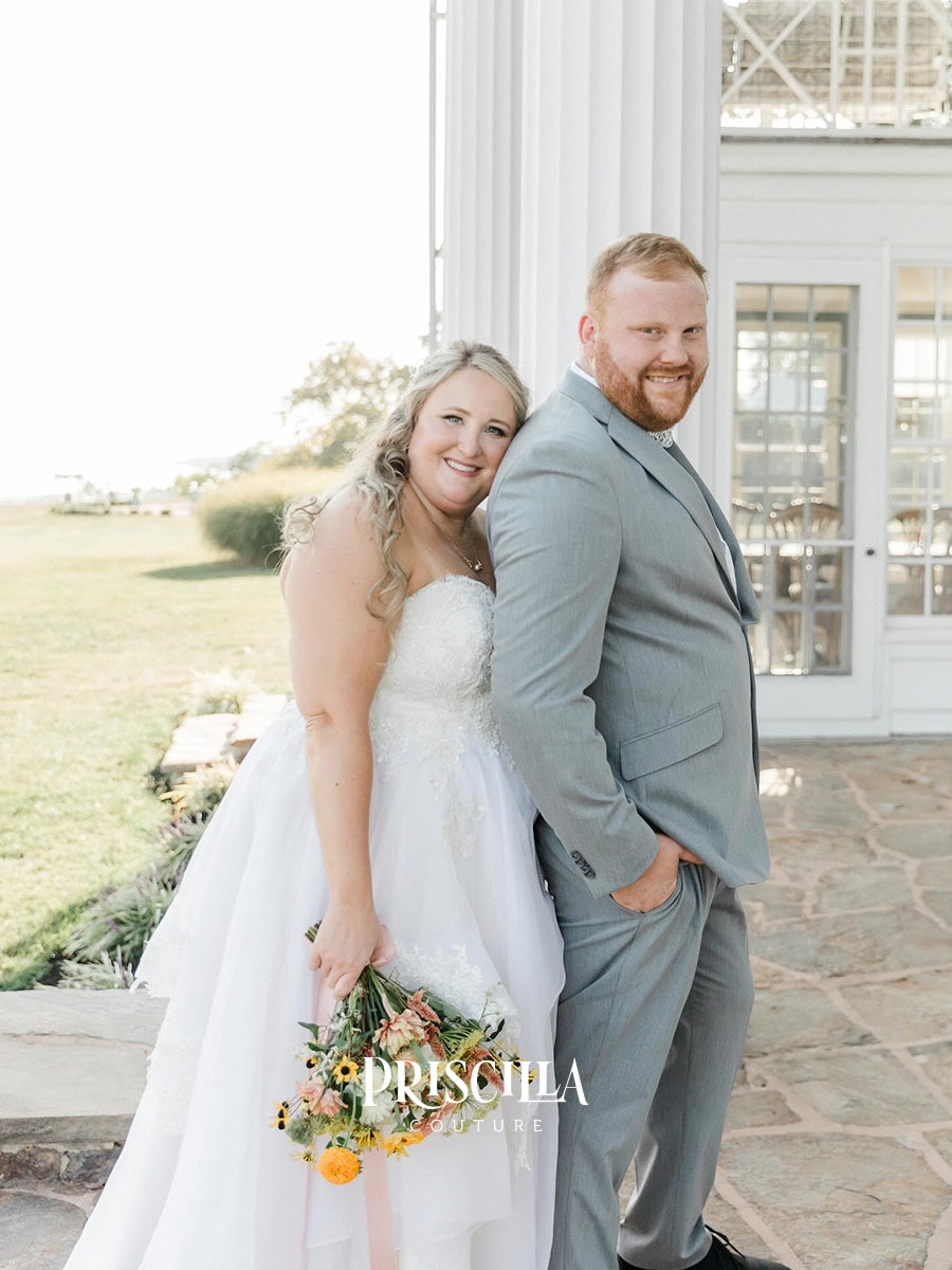 A bride in a Priscilla Couture custom white strapless wedding gown holding a floral bouquet stands next to a groom in a light gray suit on a stone patio in front of a white building. The brand name 'PRISCILLA COUTURE' is watermarked at the bottom.