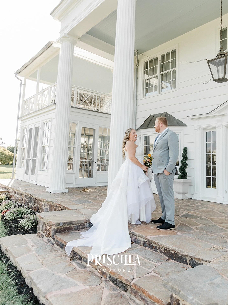 A bride in a Priscilla Couture custom long white wedding gown and a groom in a light gray suit stand on the stone steps of a large white plantation-style house porch. The bride holds a bouquet, and a watermark reading PRISCILLA COUTURE is visible near the bottom.