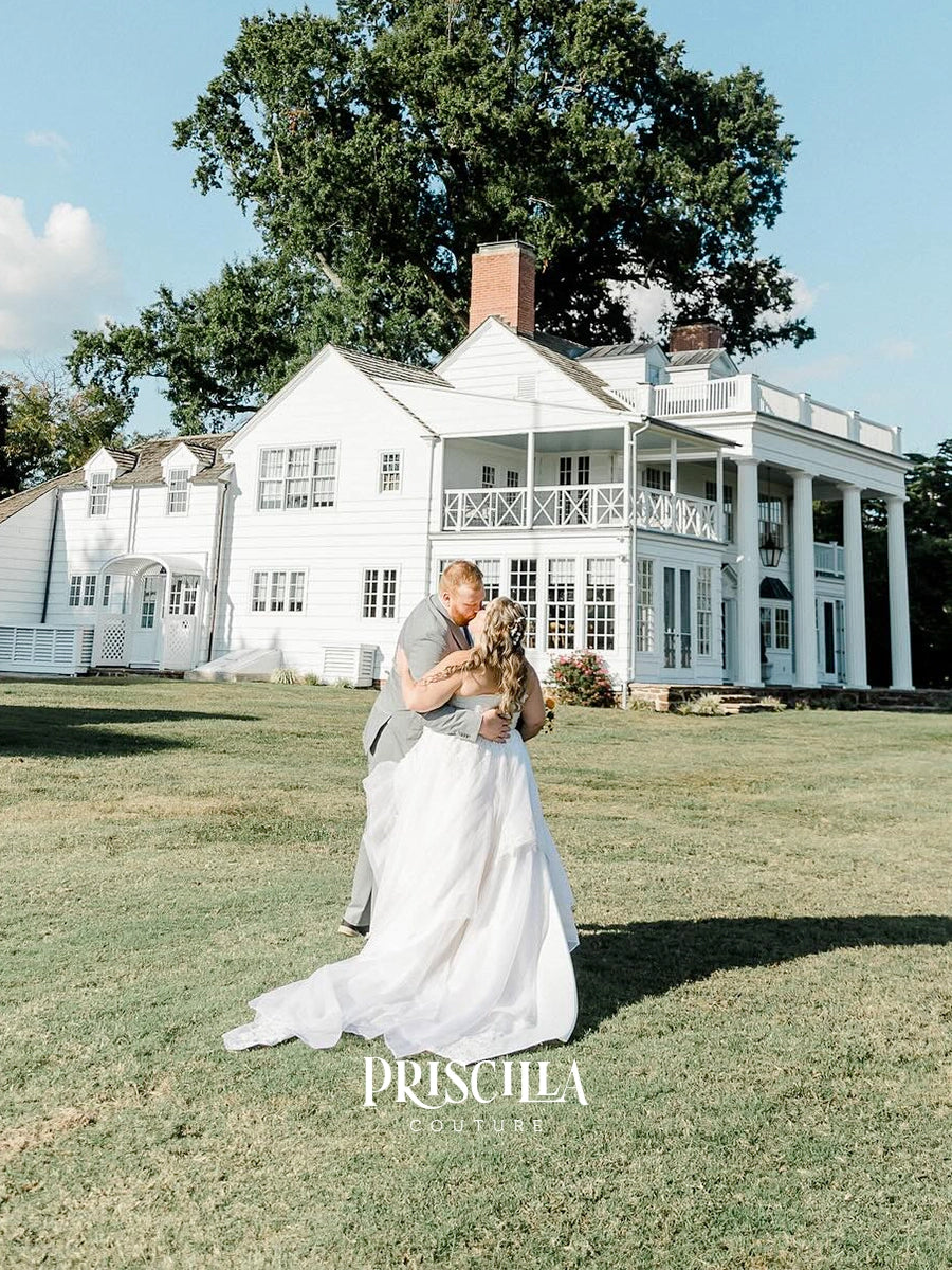 A bride in a Priscilla Couture custom white wedding gown and a man in a gray suit embrace on a grassy lawn in front of a large white plantation-style house, with a tree and blue sky behind them. The text 'PRISCHILA COUTURE' appears at the bottom.
