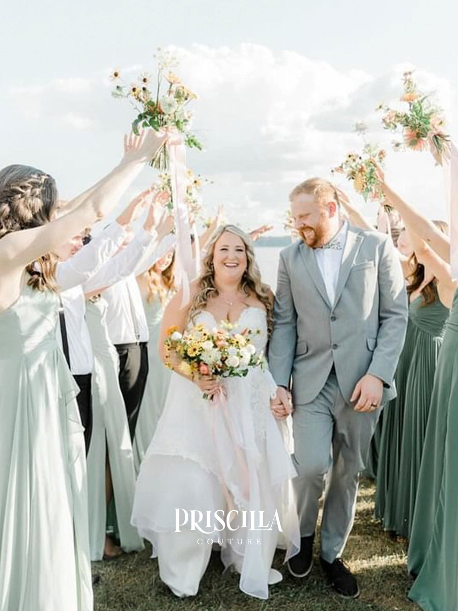 A bride in a Priscilla Couture custom white lace wedding gown and a groom in a light gray suit walk hand in hand outdoors, surrounded by bridesmaids in sage green dresses who are raising flower bouquets. The photo is taken in daylight with a cloudy sky in the background, and the brand name 'PRISCILLA COUTURE' is watermarked at the bottom.
