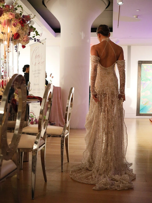 Woman in a sparkling evening gown standing in an elegant indoor setting with floral decorations.