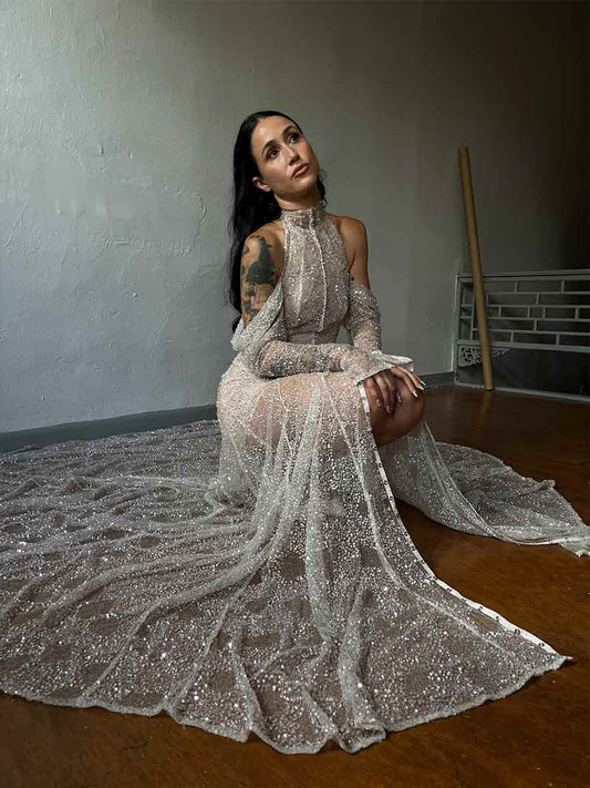 A woman seated, wearing a Priscilla Couture high neck, long sleeve, floor-length beaded wedding dress with a low back and a train.