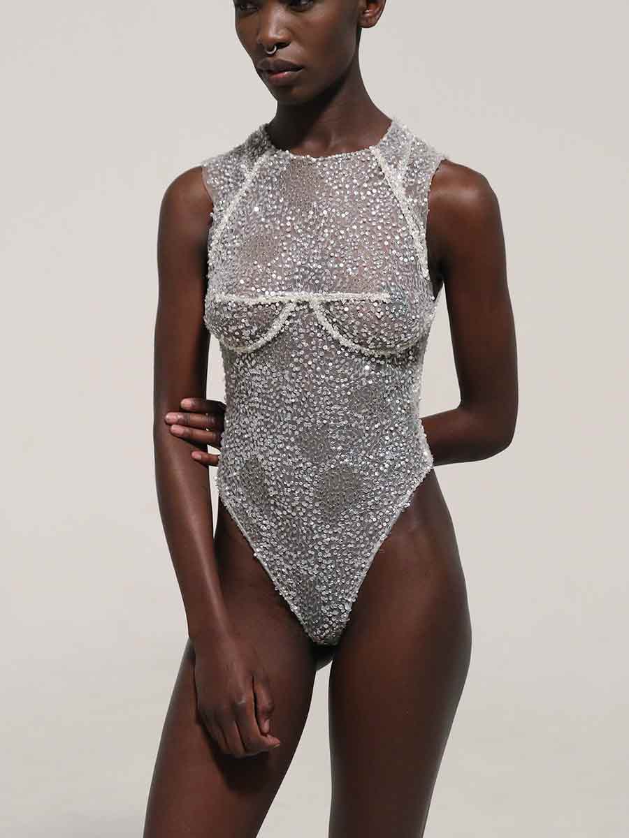 A model wearing a Priscilla Couture  sheer silver-beaded bodysuit with structured cups and a high neckline, photographed against a plain light gray studio background.