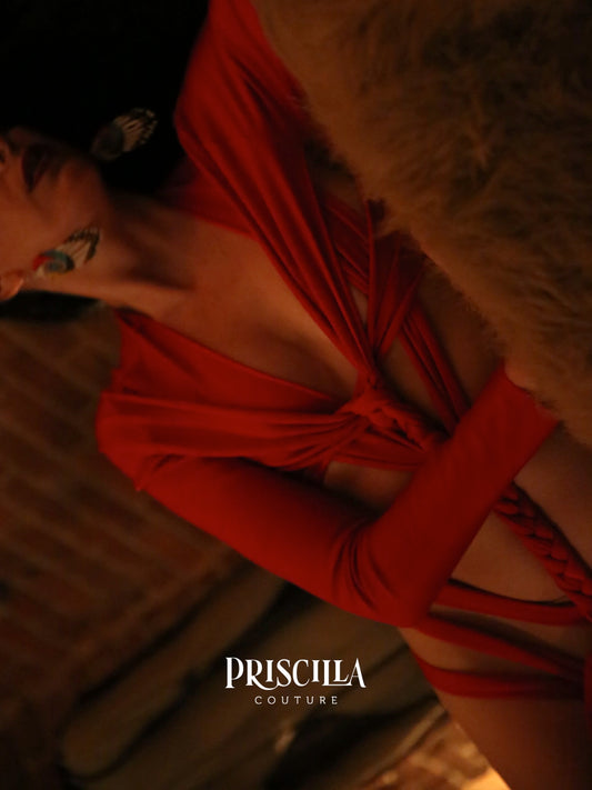 A close-up lifestyle shot of a model wearing a vibrant red long-sleeve lingerie with a twisted front detail, styled with a brown fur accessory. The brand name 'PRISCILLA COUTURE' is visible as a watermark at the bottom.