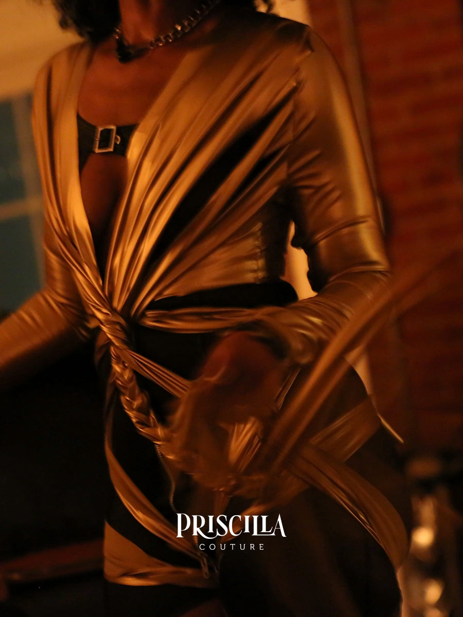 A close-up of a woman wearing a metallic gold draped wrap-style top with long sleeves and a black underlayer visible at the chest, in warm dim lighting. The brand name PRISCILLA COUTURE appears at the bottom.
